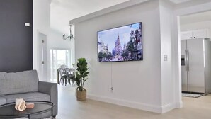 Een moderne woonkamer met een flatscreen televisie aan de muur, een grijze bank, een klein bijzettafeltje en een potplant.