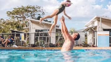 Una piscina con una persona saltando al agua, otra persona de pie en el borde, y un edificio al fondo.