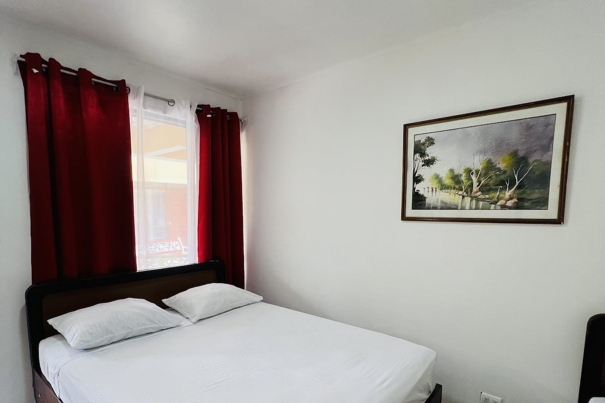 Un lit simple avec une literie blanche, des rideaux rouges, un luminaire au plafond et un tableau représentant un paysage, encadré, accroché au mur.