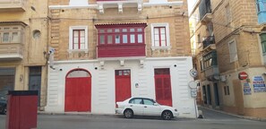A building with red shutters, a white door, and a parked white car. - Traditional Corner Townhouse for rent. Ideal for Groups or Families! (Bormla)