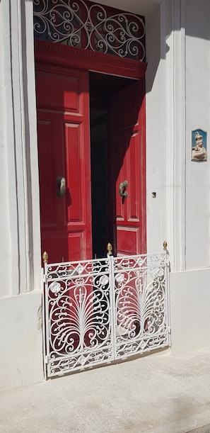 A red door with ornate metalwork, a white wall, and a small plaque.