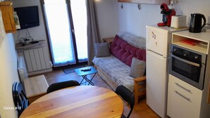 A compact living space with a sofa, a small table, a kitchenette with a fridge and oven, a TV mounted on the wall, and a window with curtains.