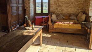 A rustic room with a stone wall, wooden furniture, a window with a view, and a table with fruits and a bottle. - Le Domaine de Magnagues (Carennac)