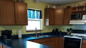 A kitchen with wooden cabinets, a blue countertop, a stove, a microwave, and a window with blue curtains. - Blue Elephant Cottage (Sullivan)