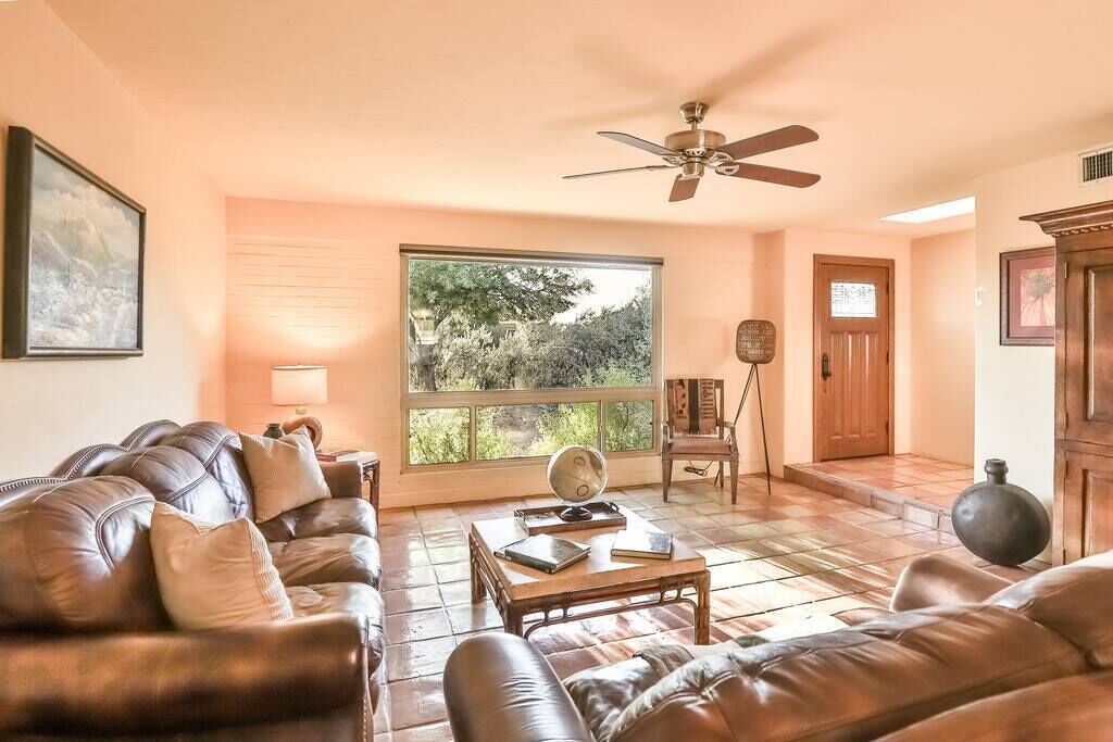 Un salon avec un canapé en cuir marron, une table basse en bois, un ventilateur de plafond, une grande fenêtre donnant sur des arbres et une porte en bois.