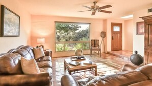 Una sala de estar con un sofá de cuero marrón, una mesa de centro de madera, un ventilador de techo, un ventanal con vistas a árboles y una puerta de madera.
