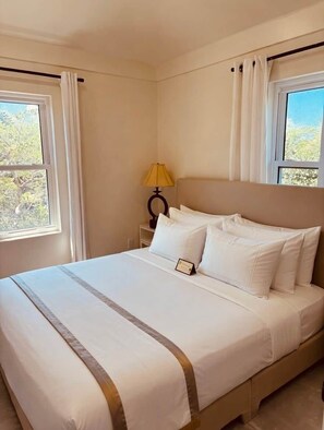 A neatly made bed with white linens and a brown accent, a bedside lamp, and a window with curtains. - Lo key cottages estate (Green turtle cay)
