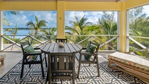 Un área de estar al aire libre techada, con una mesa, sillas y un banco, y vistas a palmeras y agua.