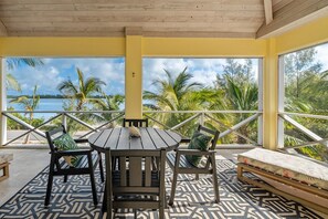 Un área de estar al aire libre techada, con una mesa, sillas y un banco, y vistas a palmeras y agua.