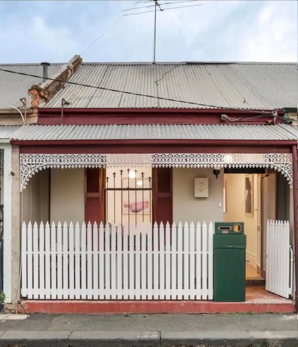 A single-story building with a corrugated metal roof, a white picket fence, and a green utility box. - A Beautiful Terrace House With Parking & Courtyard (North Melbourne)