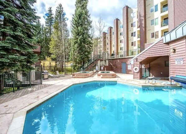 Ein Schwimmbecken, umgeben von einer Holzterrasse, mit einem Gebäude im Hintergrund.