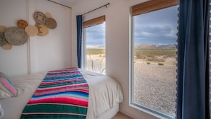 Ein Schlafzimmer mit einem Bett, einer farbenfrohen Decke, einer Wand mit gewebten Körben und Blick auf eine Wüstenlandschaft durch ein Fenster.