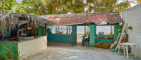 Una pequeña tienda rústica con techo de paja y fachada verde, ubicada en un entorno tropical con palmeras y vista al mar.