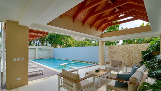 A covered outdoor pool area with lounge chairs and a table, surrounded by stone walls and tropical plants.