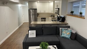 Eine moderne Küche mit Edelstahlgeräten, ein graues Sofa mit Zierkissen und ein kleines Fenster mit Jalousien.