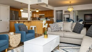 Un salon moderne avec un canapé, une table basse et des fauteuils bleus. On aperçoit, au fond, une cuisine avec des meubles en bois et un réfrigérateur.