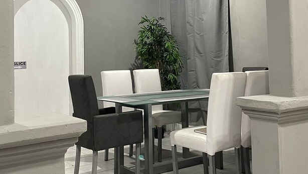 A dining area with a glass table, white chairs, a potted plant, and a clock on the wall.