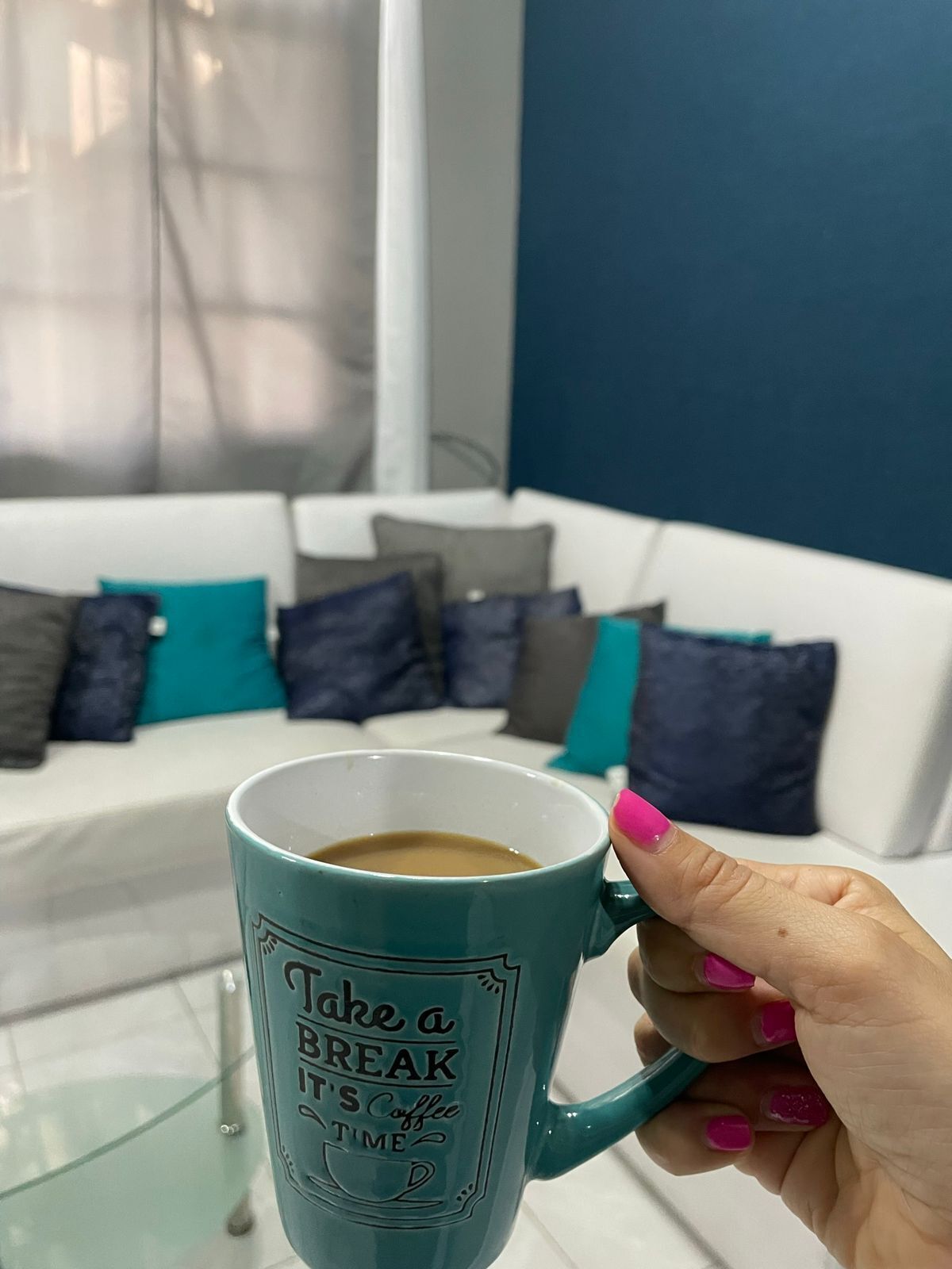 Uma caneca azul-esverdeada com uma frase motivacional, segurada por uma mão com esmalte rosa, num quarto com um sofá branco e almofadas azul-esverdeadas.