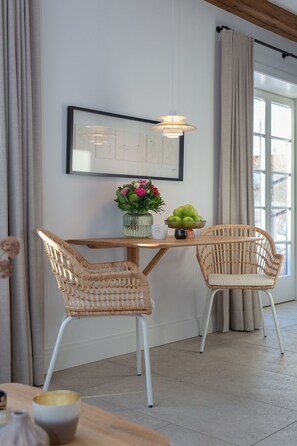 A dining area with a wooden table, two wicker chairs, a vase of flowers, and a framed picture on the wall.