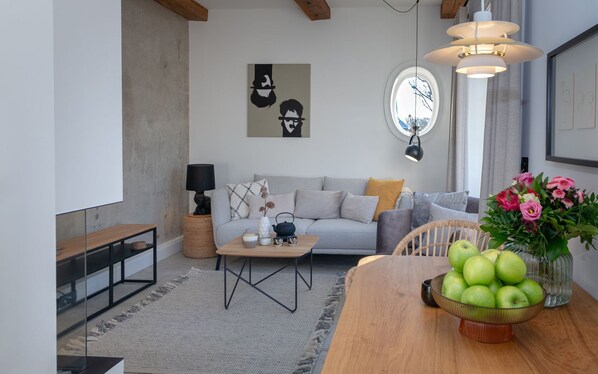 Una sala moderna con un sofá, una mesa de centro y una mesa de comedor con manzanas. Hay una lámpara, un florero con flores y una obra de arte en la pared.