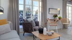 A modern living room with a grey sofa, a dark armchair, a wooden coffee table, and a dining area with wicker chairs. - NEU 2023 - Sylter Meerwind - NEU 2023 - Helle Zwei Zimmer Wohnung im Herzen von Alt Westerland (Sylt)