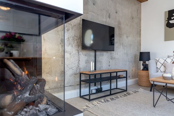 A modern living room with a fireplace, a flat-screen TV mounted on a concrete wall, a wooden coffee table, and a grey sofa.