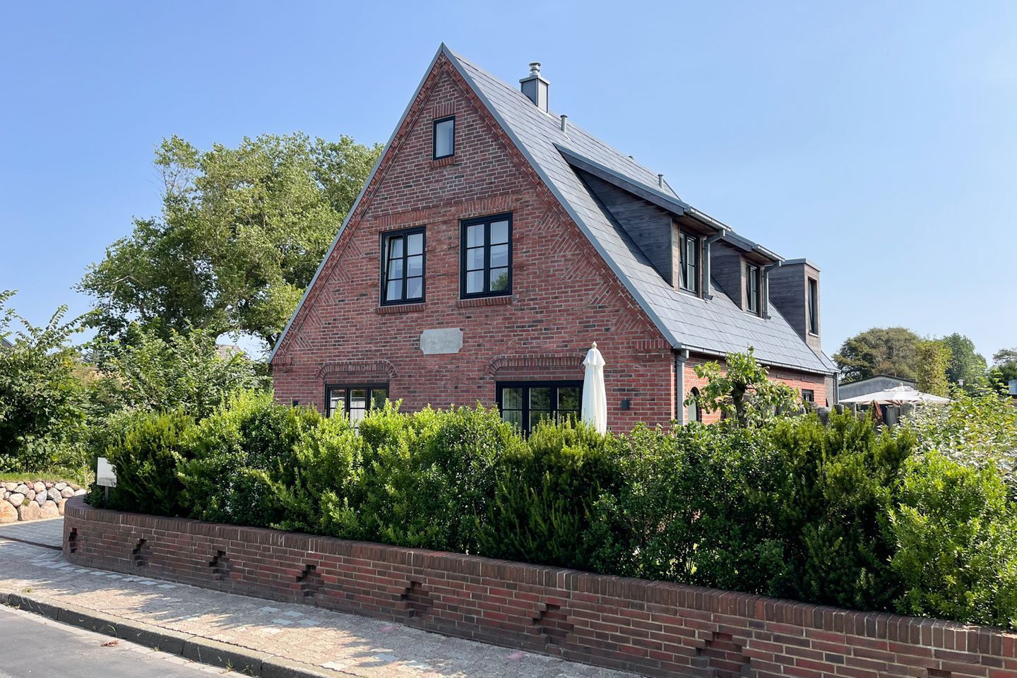 Une maison à deux étages, construite en briques, avec un toit à pignon abrupt, un petit balcon et un mur de briques surmonté d’une haie basse.