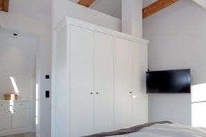 Un dormitorio con un armario blanco, una cama con una manta, un televisor en la pared y una viga de techo de madera.