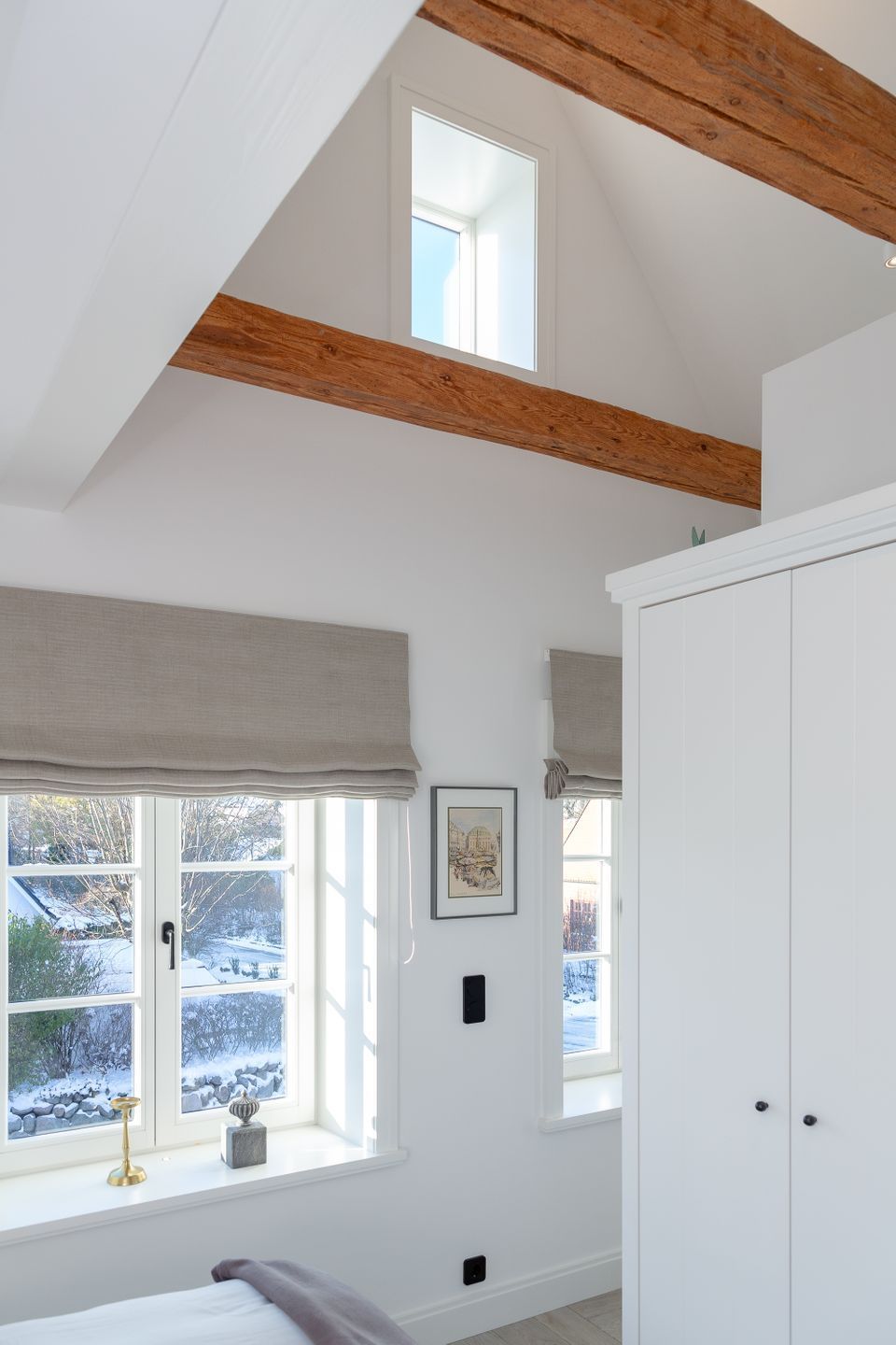Une chambre avec un plafond en pente, des poutres en bois, une grande fenêtre donnant sur un paysage enneigé, une armoire et une table de chevet avec une lampe.