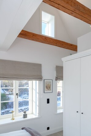 A bedroom with a sloped ceiling, wooden beams, a large window with a view of a snowy landscape, a wardrobe, and a bedside table with a lamp.
