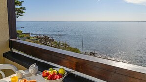 A breakfast table with croissants, fruit, and juice, overlooking a calm sea with a clear sky.