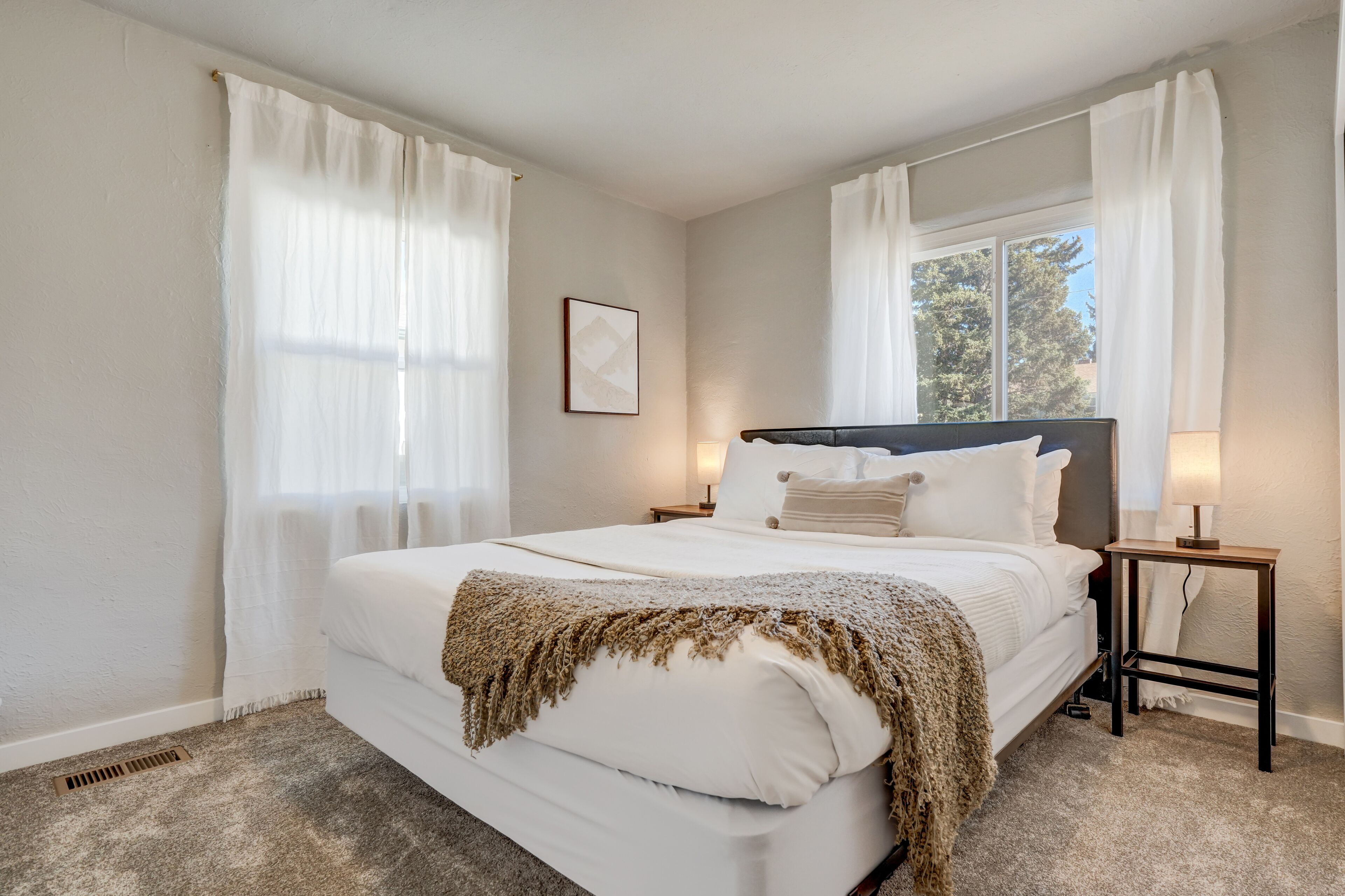 Un dormitorio con una cama grande, cortinas blancas, una mesita de noche con una lámpara y un cuadro enmarcado en la pared.