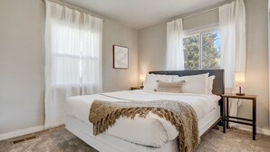 A bedroom with a large bed, white curtains, a bedside table with a lamp, and a framed picture on the wall. - Bon Voyage (Colorado Springs)
