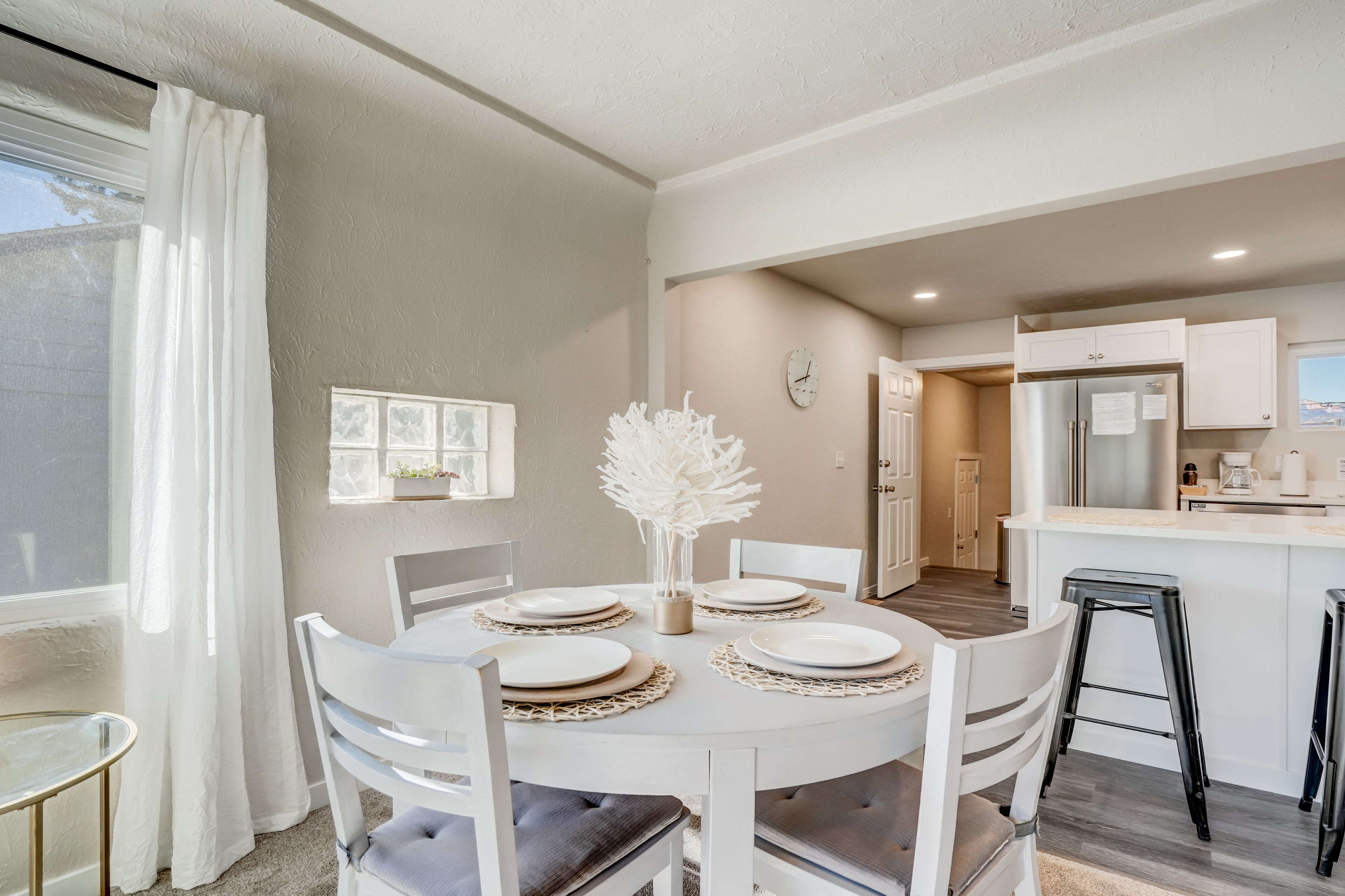 Un comedor con una mesa redonda dispuesta para cuatro personas, sillas blancas y un jarrón con flores blancas. Se ve una cocina al fondo, con un refrigerador y armarios.