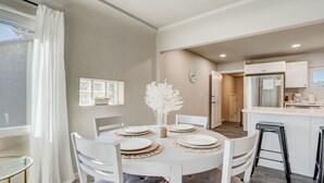 A dining area with a round table set for four, white chairs, and a vase of white flowers. A kitchen is visible in the background with a refrigerator and cabinets. - Bon Voyage (Colorado Springs)