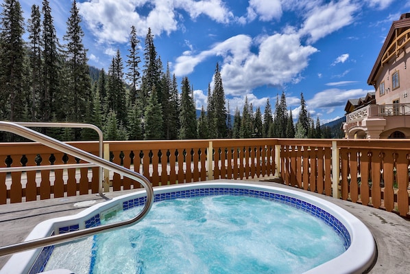 A hot tub with a wooden deck and a railing, surrounded by a forest and a mountain in the background. - Hearthstone Lodge #421 (Sun Peaks)