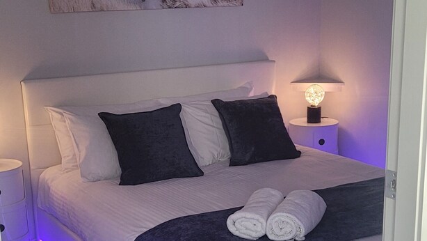 Une chambre à coucher avec un lit, des oreillers, une table de chevet, une lampe fixée au mur et un tableau.