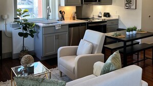 A modern kitchen with grey cabinets, a stainless steel refrigerator, and a microwave. There is a dining table with chairs and a small potted plant on the windowsill. - Serene Urban Oasis (Louisville)