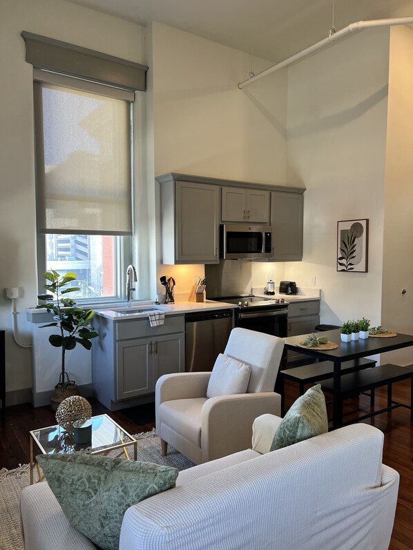 A modern kitchen with grey cabinets, a stainless steel refrigerator, and a microwave. There is a dining table with chairs and a small potted plant on the windowsill. - Serene Urban Oasis (Louisville)