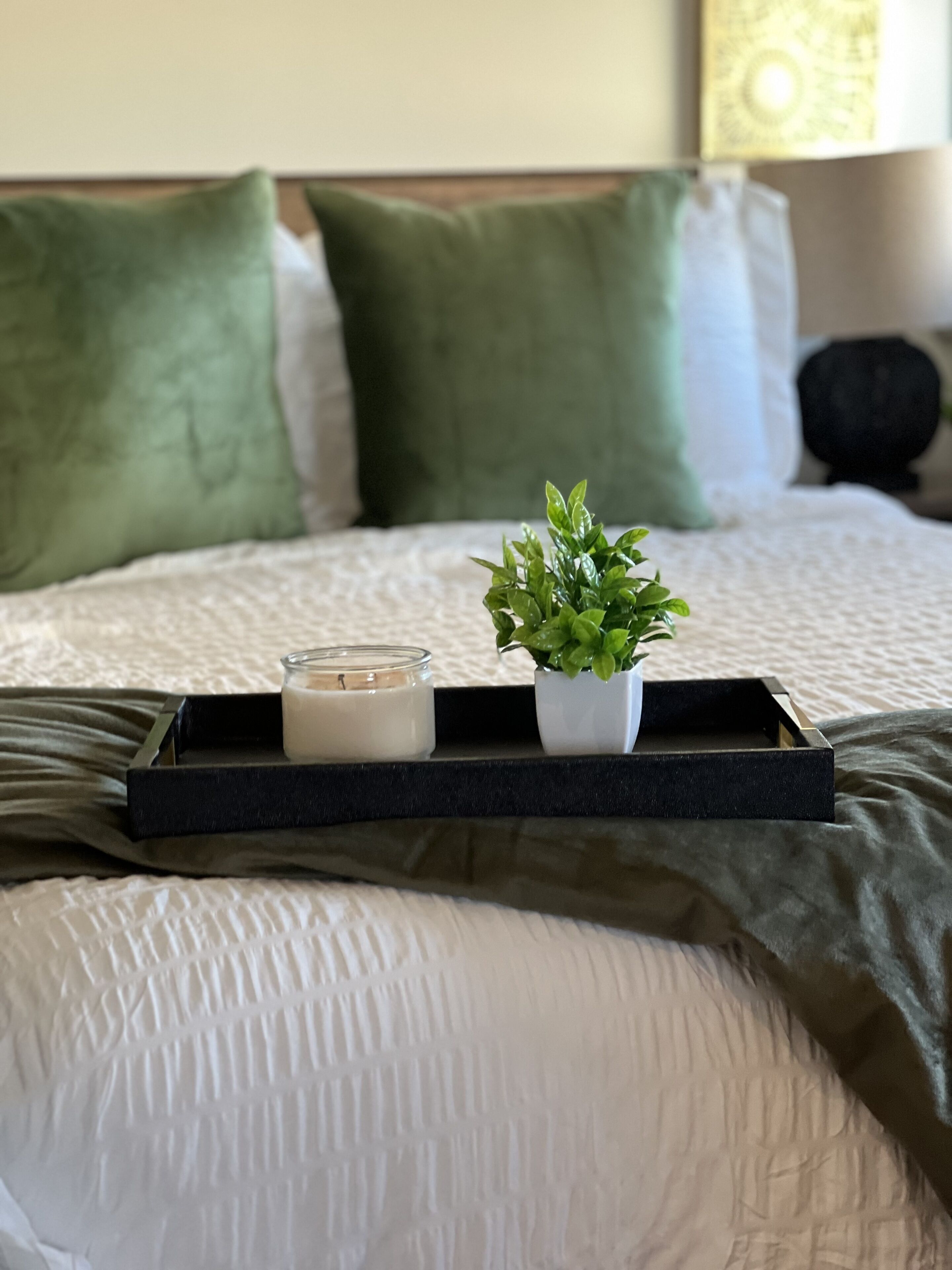 A neatly made bed with a white comforter, green pillows, a small potted plant, and a candle on a tray.