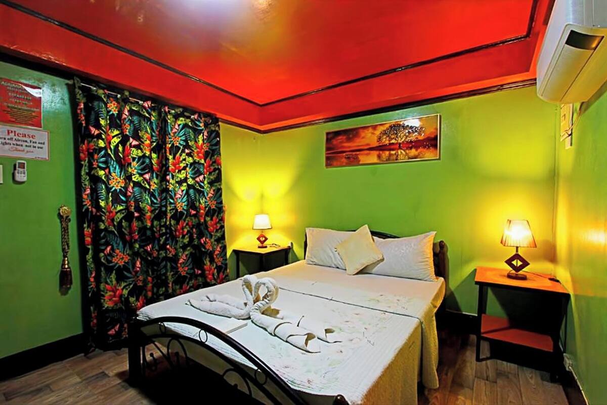 A hotel room with a bed, two bedside tables with lamps, a floral curtain, a painting on the wall, and a sign on the door.