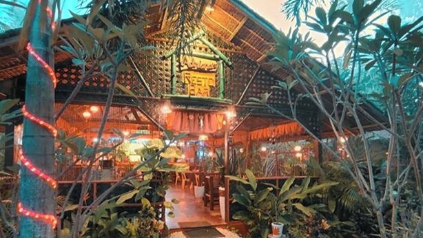 Restaurante com temática tropical, com telhado de palha e rodeado por vegetação exuberante.