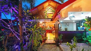 Um hotel com entrada em um jardim tropical, fachada de madeira e uma área de recepção visível.