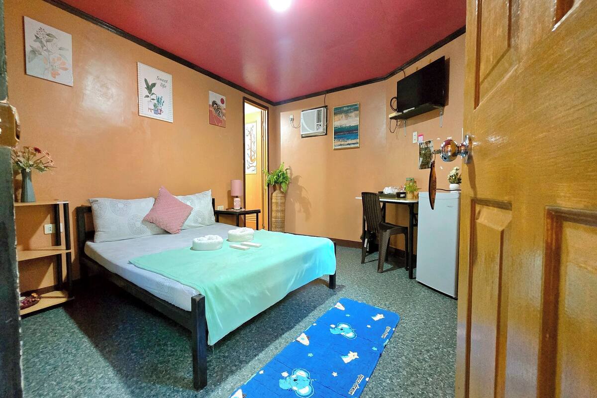 A bedroom with a bed, a desk, a refrigerator, and a TV on the wall.