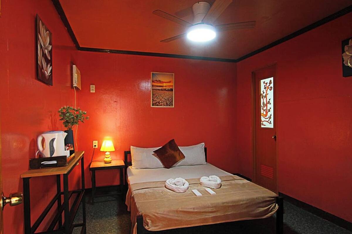 A hotel room with a red wall, a bed with white linens, a ceiling fan, a nightstand with a lamp, and a painting on the wall.