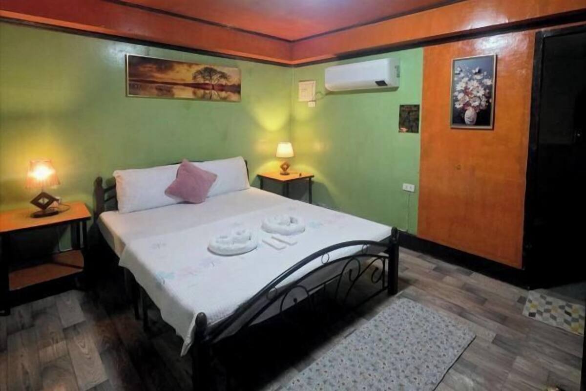A single bed with pillows and a blanket, a nightstand with a lamp, a wooden headboard, and a wall-mounted air conditioner.