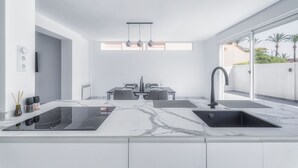 Eine moderne Küche mit einer zentralen Kochinsel, einem Essbereich mit Tisch und Stühlen und einem großen Fenster mit Blick auf Palmen.
