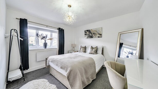Une chambre à coucher comprenant un lit, des tables de chevet, une commode, un miroir, une fenêtre avec des rideaux et un luminaire suspendu.
