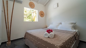 Ein ordentlich bezogenes Bett mit einer braunen Tagesdecke und weißen Kissen, ein Fenster mit Blick ins Grüne und eine Bambuspflanze neben dem Bett.