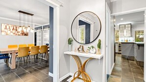 A modern dining area with a wooden table and yellow chairs, a console table with a mirror and decorative items, and a kitchen visible through an open door. - 5 BR w/pool, minutes to Six flags, AT&T stadium (Grand Prairie)